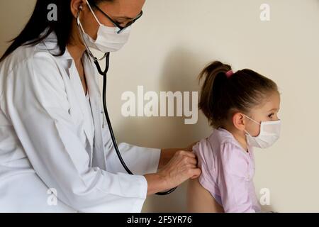 chubby bambina in esame pediatrico da parte del medico, che ascolta i polmoni e il cuore e indossare maschere protettive per il viso Foto Stock