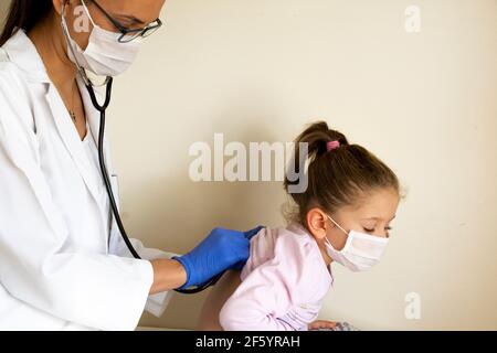 chubby bambina in esame pediatrico da parte del medico, che ascolta i polmoni e il cuore e indossare maschere protettive per il viso Foto Stock