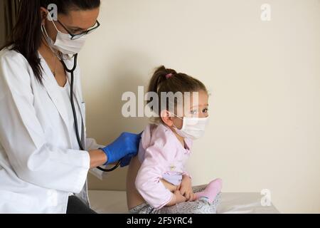 chubby bambina in esame pediatrico da parte del medico, che ascolta i polmoni e il cuore e indossare maschere protettive per il viso Foto Stock