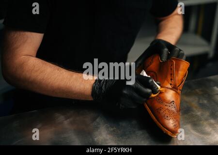 Mani ravvicinato di irriconoscibile calzolaio in guanti neri che sfregano la vernice su scarpe in pelle marrone chiaro con le dita. Foto Stock