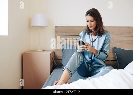 Bella giovane donna in pigiama di seta seduta sul letto e. rispondere alle domande sullo smartphone Foto Stock