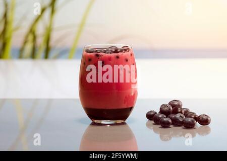 Primo piano di un bicchiere di succo di frutta con mirtilli accanto ad altri mirtilli su un tavolo di vetro su uno sfondo sfocato. Vegano e sano Foto Stock