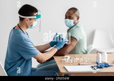 Giovane medico in maschera protettiva e uniforme dà iniezione shot al paziente afroamericano Foto Stock