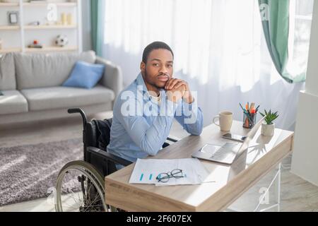 Lavoro online per disabili. Serio ragazzo nero in sedia a rotelle seduto alla scrivania con il computer portatile, guardando la fotocamera, in interni Foto Stock