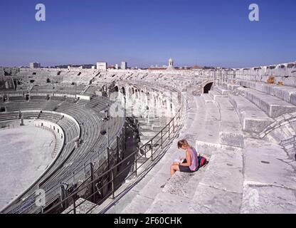 Historical1990 archivio vista interna della preservata Arena francese di Nîmes un famoso anfiteatro romano visto nel 1993 dal livello più alto con giovane turista donna seduta sopra terrazza in pietra con vista interna degli anni '90 fino al funzionamento eventi arena e i livelli più bassi di posti a sedere su un caldo giorno del sole del cielo blu in un'immagine di viaggio archivistica in Il dipartimento del Gard della Regione Occitanie della Francia meridionale Foto Stock