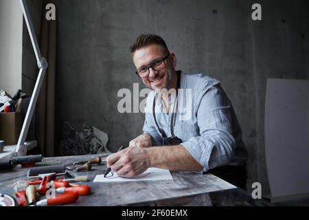 Artista maschile bearded di mezza età in occhiali seduti su uno sfondo di lavoro. Maestro di linocut in grembiule sorridente e guardando dritto in macchina fotografica. Foto di alta qualità Foto Stock