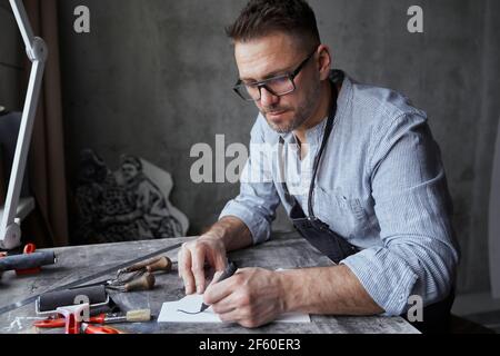 Bell'uomo caucasico bearded in grembiule e bicchieri seduti al tavolo in studio facendo schizzo con pennarello. Design professionale o di design. Foto di alta qualità Foto Stock