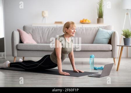 Yoga domestico. Donna matura flessibile che fa la cobra asana su tappetino sportivo, l'addestramento al tutorial online su laptop, in interni Foto Stock