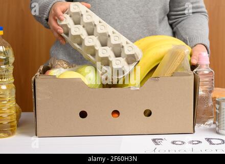 donna in guanti continua a raccogliere cibo, frutta e cose e una scatola di cartone per aiutare coloro che hanno bisogno, il concetto di aiuto e volontariato Foto Stock