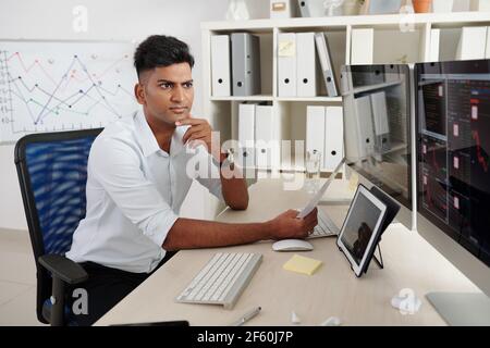 Il giovane operatore indiano accigliato che controlla il mercato azionario che guarda schermate che analizzano il prezzo delle scorte Foto Stock