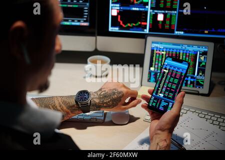 Trader che lavora a tarda notte e controlla i dati dei mercati azionari tramite applicazione sullo smartphone Foto Stock