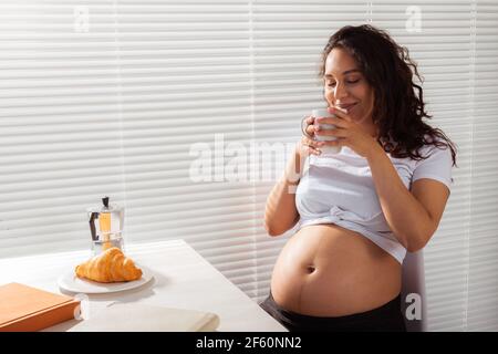 Gioiosa giovane incinta bella donna guarda attraverso le persiane durante la sua colazione mattutina con caffè e croissant. Concetto di Buongiorno e. Foto Stock