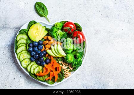 Concetto di dieta sana del cuore. Insalata con verdure, olio d'oliva, frutti di bosco e noci in un piatto a cuore, vista dall'alto. Foto Stock