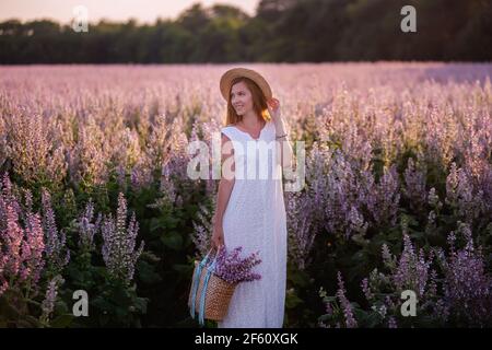 Una giovane donna in una terrina bianca, cappello di paglia tiene un cesto di vimini con un bouquet. Una ragazza cammina attraverso un campo rosa in fiore salvia al tramonto. Il conc Foto Stock