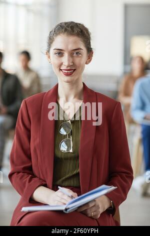 Ritratto verticale della giovane donna d'affari di successo che indossa rosso e sorridente con la fotocamera mentre si è seduti sulla sedia in ufficio Foto Stock
