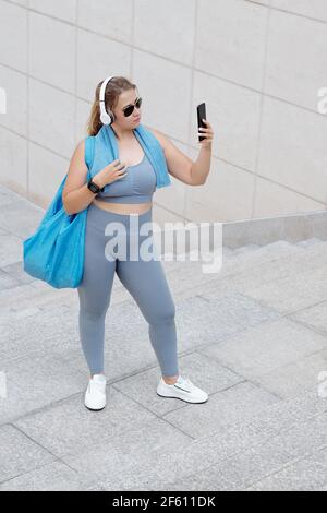 Bella fit Plus taglia giovane donna con palestra in posa borsa per selfie dopo l'allenamento in occhiali da sole e cuffie Foto Stock