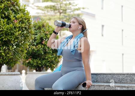 Sete stanco più taglia giovane donna che beve acqua dopo intenso formazione all'aperto Foto Stock