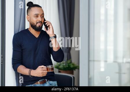 Ritratto di uomo bearded positivo che beve il caffè del mattino e che parla al telefono con un collega o un dipendente Foto Stock