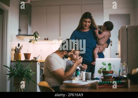 Padre che alimenta il figlio mentre la madre sta con la bambina dentro cucina Foto Stock