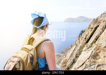 Giovane ragazza turistica con uno zaino si erge sulla cima della scogliera e guarda al mare. Foto Stock