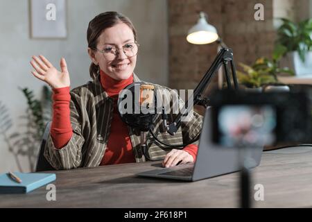 Giovane donna in occhiali seduto al tavolo con microfono e il laptop e la posa alla fotocamera durante la registrazione Foto Stock