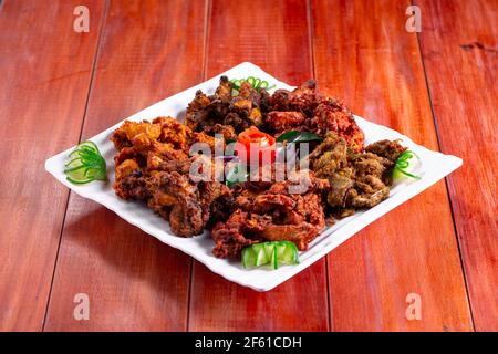 Pakoda di pollo o pakora, tre diversi sapori di pakora disposti in un piatto bianco quadrato e guarnito con pomodoro e cetriolo su un dorso di legno Foto Stock