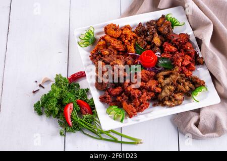 Pakoda di pollo o pakora, tre diversi sapori di pakora disposti in un piatto bianco quadrato e guarnito con pomodoro e cetriolo su uno sfondo bianco Foto Stock