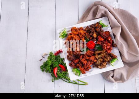 Pakoda di pollo o pakora, tre diversi sapori di pakora disposti in un piatto bianco quadrato e guarnito con pomodoro e cetriolo su uno sfondo bianco Foto Stock