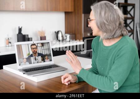 Donna matura che tiene una videoconferenza sul laptop seduto a casa, la donna anziana ha coinvolto una riunione virtuale con i colleghi, guardando un webinar video online Foto Stock