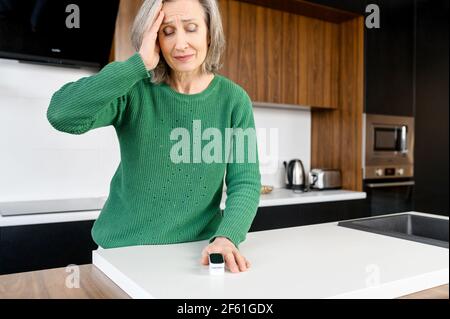 La donna con capelli grigi di media età si sente malata, misurando la pressione sanguigna e la frequenza del polso, tenendo la testa e soffre di mal di testa. Donna anziana che usa ossimetro a casa Foto Stock