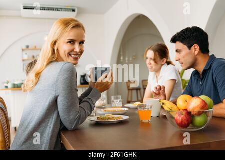 Ritratto di bella giovane madre che tiene una tazza di caffè mentre sorridendo e guardando la macchina fotografica con il marito bello e i bambini in una conversazione durante la prima colazione Foto Stock