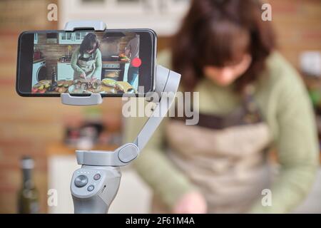 Giovane donna che cucinava melanzane ripiene e registrava un video con il suo smartphone per il suo canale di cucina. Creatore di contenuti e influencer. Foto Stock