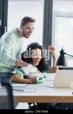 uomo d'affari allegro che punta al laptop vicino eccitato collega afroamericano mostrare il gesto di vittoria Foto Stock