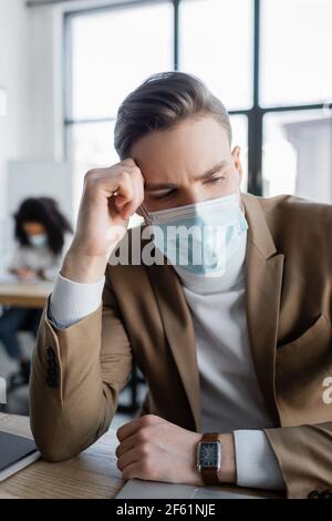 uomo d'affari sconvolto in maschera medica seduto in ufficio mentre africano collega americano che lavora su sfondo sfocato Foto Stock