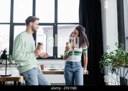 partner commerciali interrazziali felici in piedi con il caffè per andare durante conversazione in ufficio Foto Stock