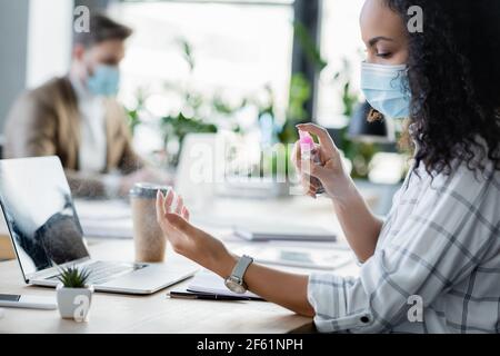 donna d'affari afroamericana che applica l'igienizzatore della mano vicino al collega che lavora sopra sfondo sfocato Foto Stock