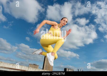 Donna sportiva che indossa abiti attivi gialli a mezz'aria, saltando durante la corsa. Foto Stock