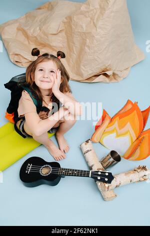 Sognante bambina seduta accanto ad un falò falso con fiamme di carta e tronchi di betulla. Sta guardando in su, appoggiandosi sulla sua mano sopra lo sfondo blu. Alta Foto Stock