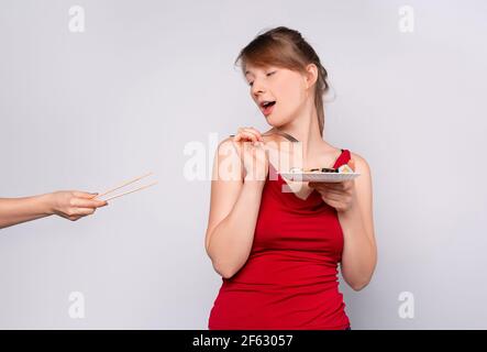 Una giovane bionda rifiuta i bastoncini di legno e preferisce una forchetta per il cibo. La ragazza mangia il sushi con una forchetta. Sfondo grigio studio. Foto Stock