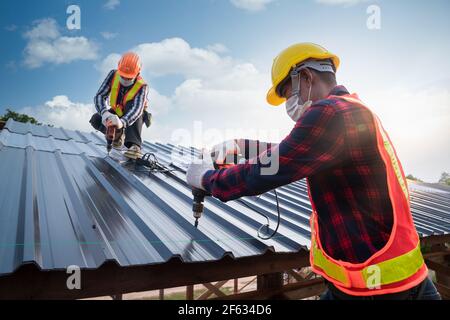 Due indumenti di sicurezza per i lavoratori Roofer che utilizzano una pistola pneumatica o pneumatica per unghie e che si installano su una nuova lamiera del tetto, concetto di tetto di edificio residenziale sotto la const Foto Stock