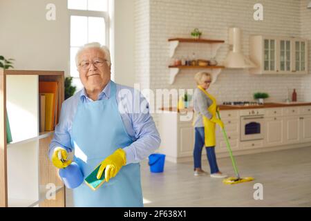L'uomo anziano pulisce i ripiani tenendo un panno e un detergente mentre sua moglie lava il pavimento. Foto Stock