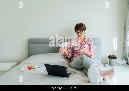 Donna emozionale in occhiali, camicie e pantaloni pigiama che hanno videochattare sul suo laptop seduto a letto. Cercate il lavoro remoto da casa Foto Stock