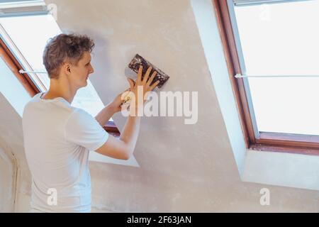 Un giovane che graffia con una spatola e rimuove la vecchia carta da parati su un muro di una stanza. Fai da te concetto. Ristrutturazione della casa. Messa a fuoco selettiva, copia Foto Stock