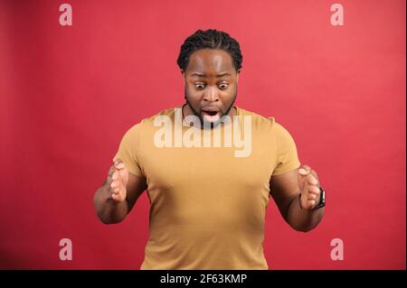 Uomo afro-americano scioccato che presenta qualcosa, bocca aperta ragazzo skinned scuro che tiene spazio vuoto per il vostro prodotto, loks giù, dimostra dimensione isolata su sfondo rosso, concetto di mockup Foto Stock