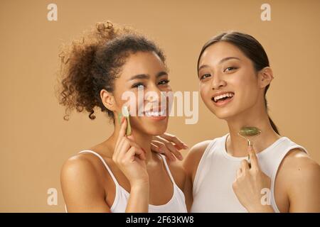 Graziose giovani donne che sorridono alla macchina fotografica, usando il rullo di giada e gua sha facciale mentre posando insieme isolato su sfondo beige Foto Stock