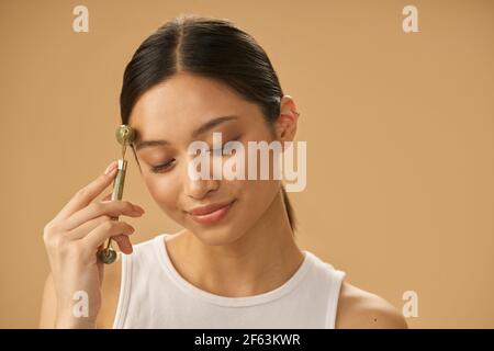 Giovane donna rilassata che usa il rullo di giada per massaggiare il viso, posando con gli occhi chiusi isolati su sfondo beige Foto Stock