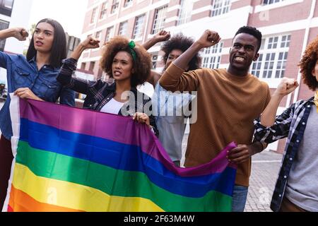 Diversi manifestanti maschili e femminili su marzo con bandiera arcobaleno, gridando e sollevando pugni Foto Stock