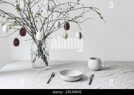 Impostazione del tavolo festivo di Pasqua. Rami freschi in vaso decorato da ornamenti di carta. Uova di pollo appesi con decorazione interna di quail festher.Spring Foto Stock