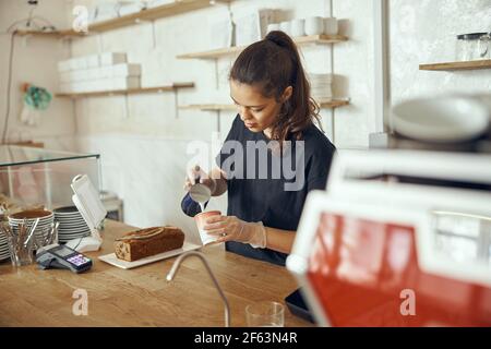 Barista giovane donna che versa latte fresco per preparare caffè latte per il cliente al bar. Per le piccole imprese startup nel concetto di industria alimentare Foto Stock