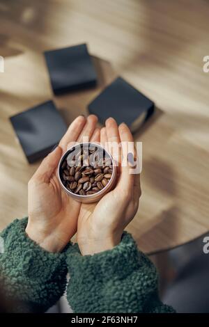 La mano femminile tiene la ciotola di caffè macinato e fagioli nella confezione regalo sul tavolo, primo piano Foto Stock